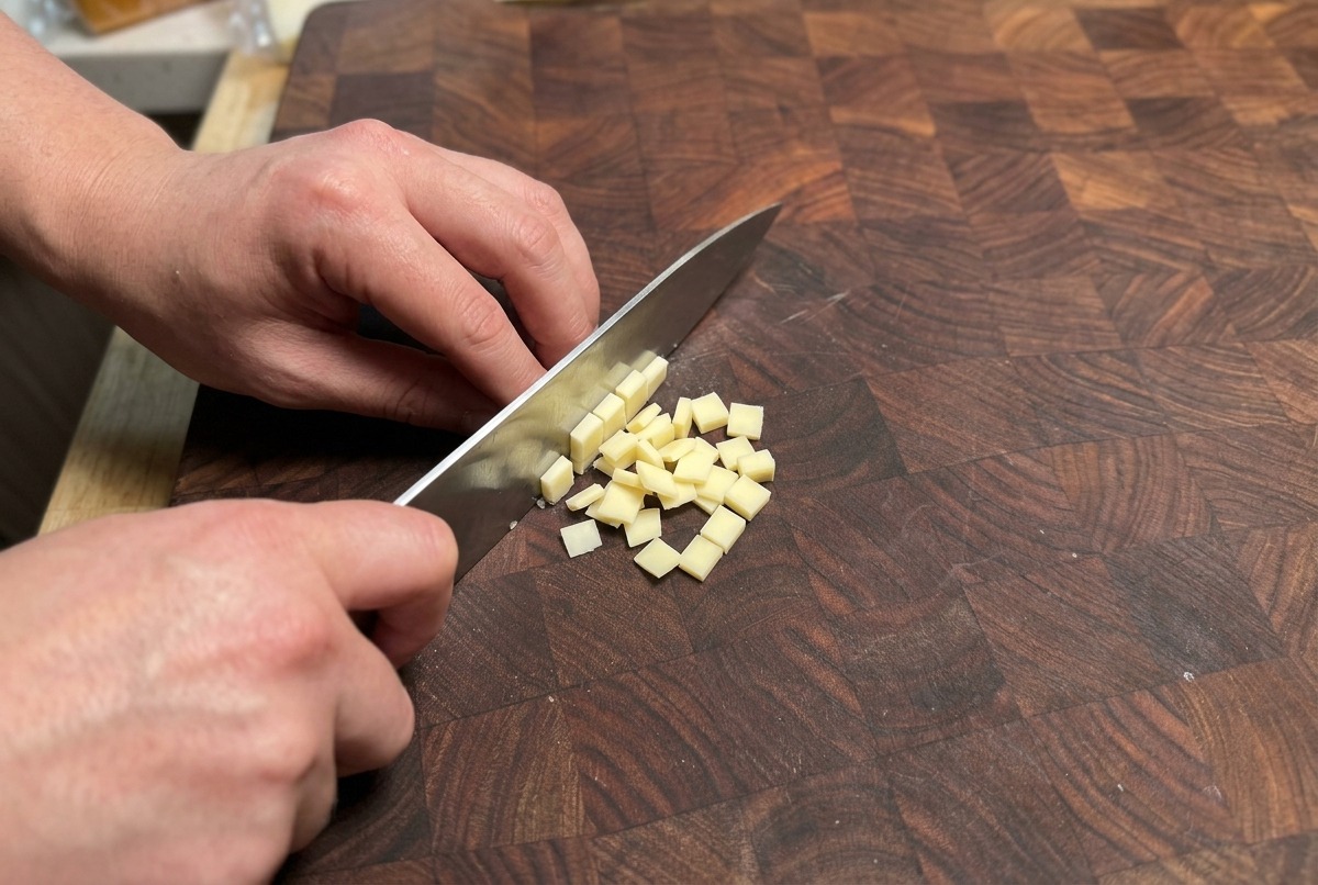 Manos usando un cuchillo de chef para cortar un bloque de queso amarillo pálido en cubos pequeños sobre una tabla de madera.