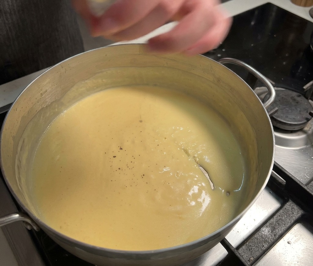 Pimienta negra fresca moliéndose sobre una salsa de queso amarilla, espesa y cremosa que hierve a fuego lento en una olla.