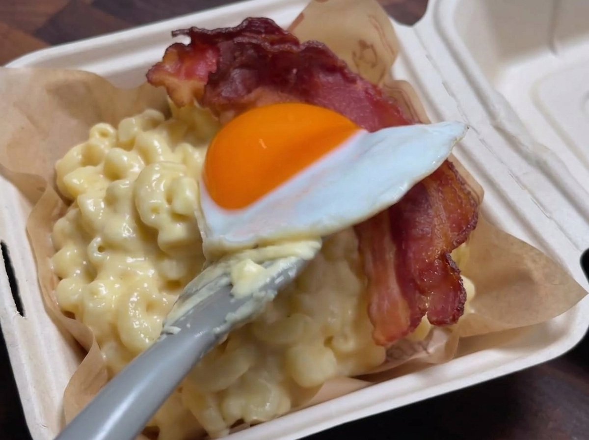 Una espátula colocando suavemente un huevo frito sobre una tira de tocino crujiente que reposa en una porción de macarrones con queso cremosos.