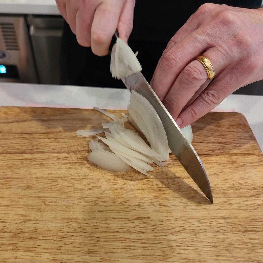 Primer plano de unas manos usando un cuchillo de chef para cortar una cebolla blanca en tiras finas sobre una tabla de madera clara.
