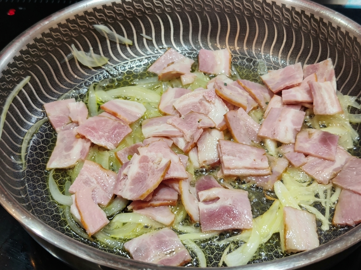 Tiras de tocino rosado friéndose en una sartén junto a tiras de cebolla tiernas y translúcidas en aceite.