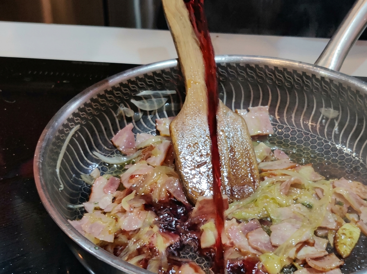Un chorro de vino tinto oscuro vertiéndose desde una copa en una sartén de acero inoxidable llena de tocino y cebollas chisporroteantes.