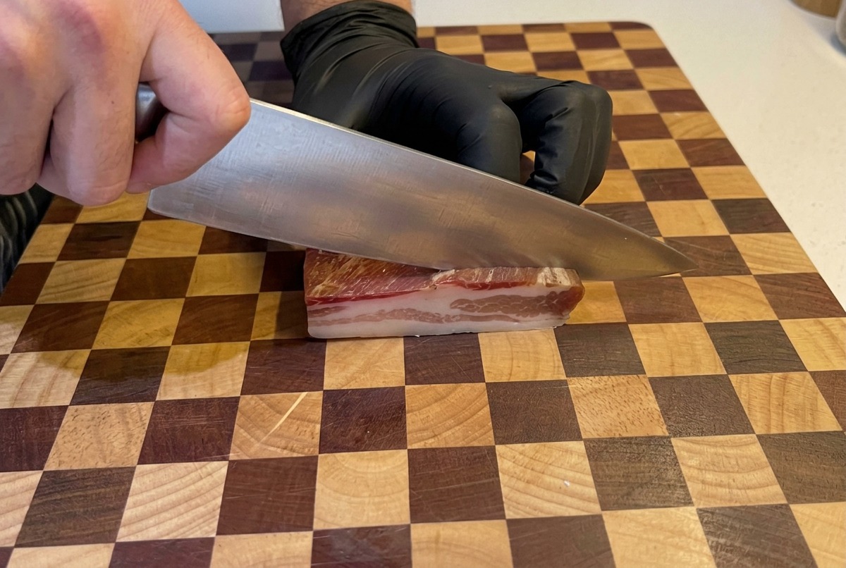 Manos con guantes negros usan un cuchillo de chef grande para cortar un trozo grueso de panceta en dados pequeños sobre una tabla de madera.