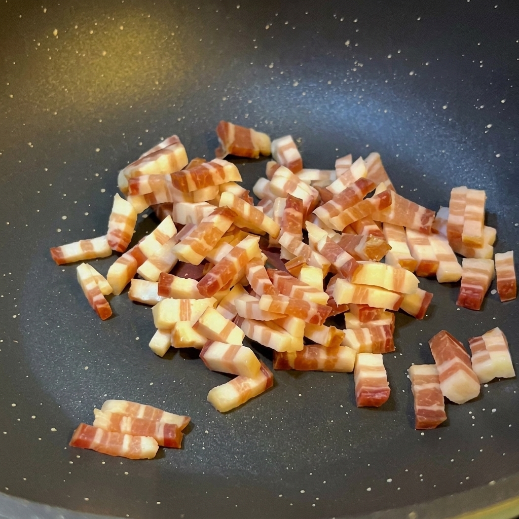 Dados pequeños de panceta italiana salteándose en una sartén antiadherente oscura mostrando la grasa derretida y un ligero dorado en los bordes.