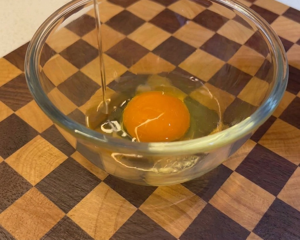 Un huevo crudo siendo cascado y vertido en un bol de cristal que ya contiene una yema de huevo con un fondo de madera.