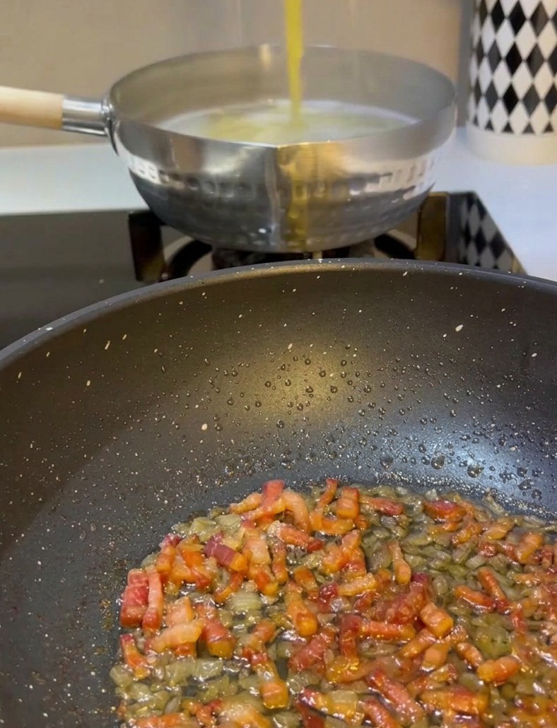 Una mano usando pinzas de metal para levantar espaguetis amarillos de una olla de agua hirviendo y pasándolos a una sartén con panceta y cebolla frita.