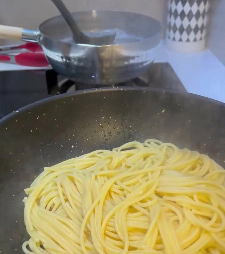 Un cucharón pasando agua turbia de una olla hirviendo de pasta a una sartén que ya contiene espaguetis y carne cocinada.
