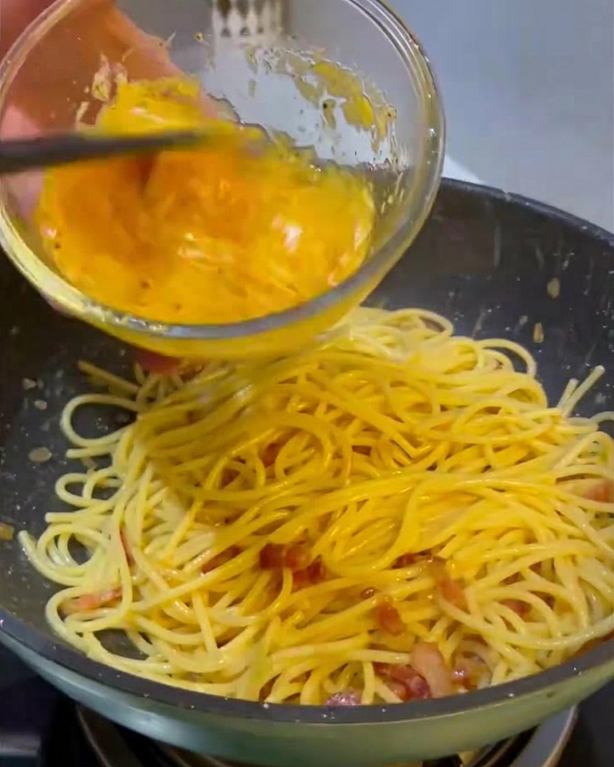 Vertiendo una mezcla amarilla vibrante de huevos y queso desde un bol de cristal sobre una sartén de espaguetis calientes y carne frita.