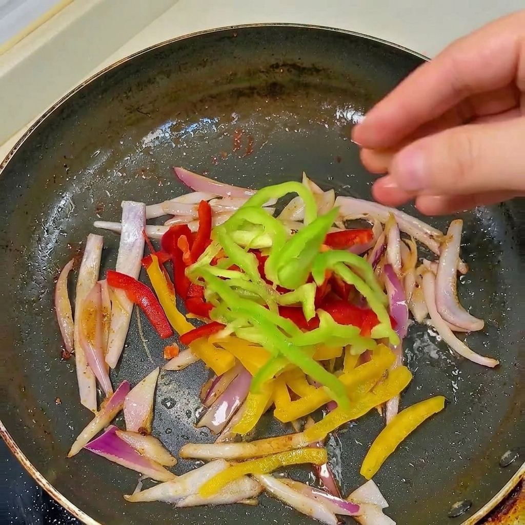 Una mano agregando tiras de pimiento rojo, amarillo y verde en una sartén negra con cebolla salteada.