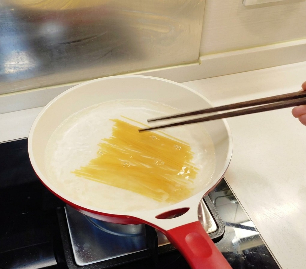 Espaguetis secos introduciéndose en una sartén blanca con agua hirviendo usando palillos de madera oscuros.