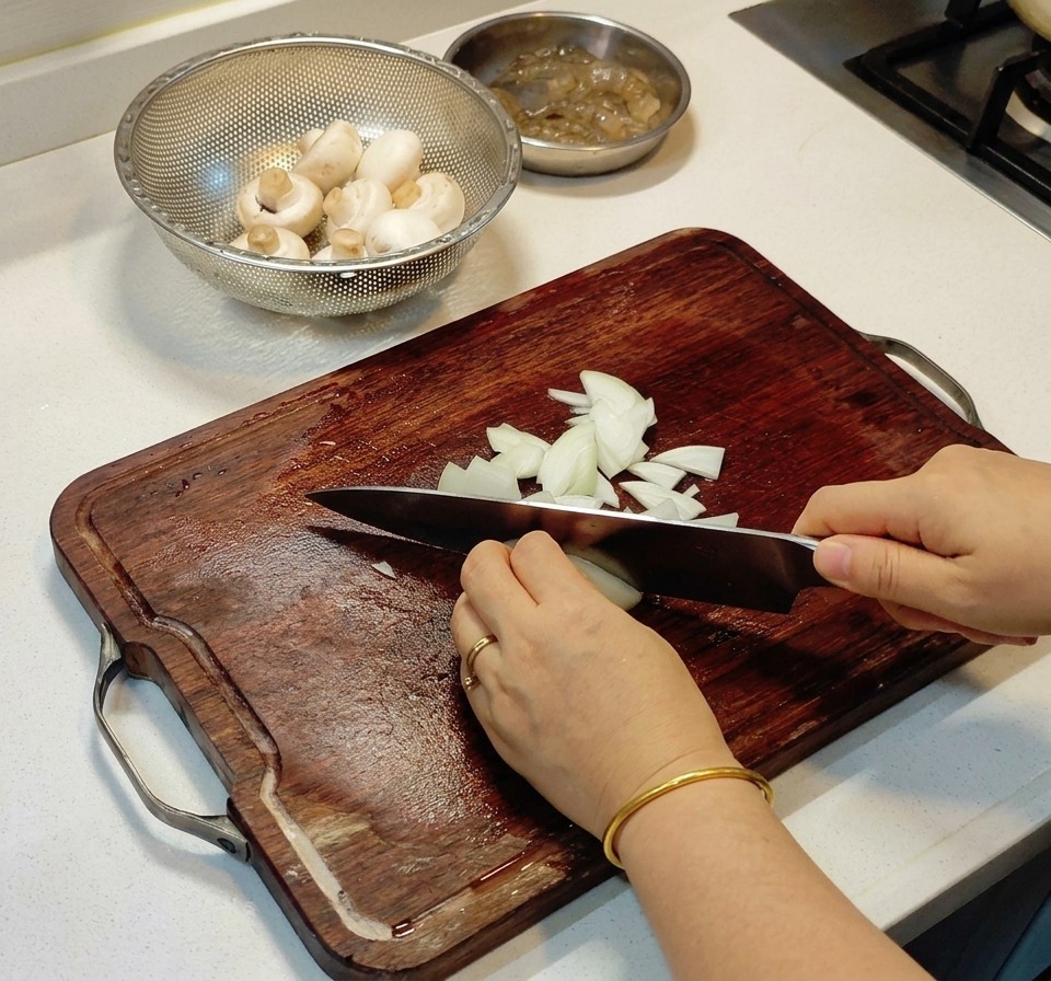 Manos usando un cuchillo de chef para picar finamente una cebolla blanca en una tabla de cortar de madera oscura junto a un cuenco con champiñones.