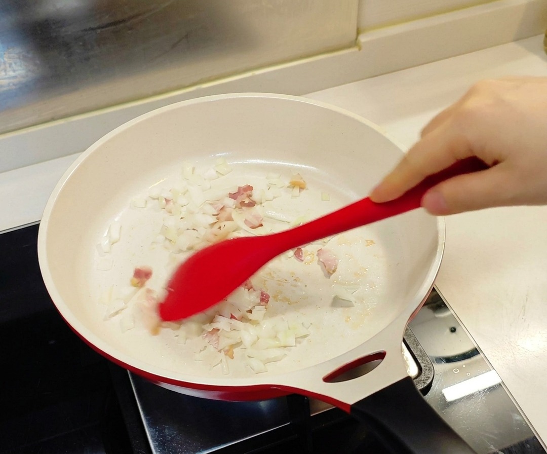 Una espátula roja removiendo cebolla picada y trozos pequeños de tocino en una sartén blanca con revestimiento cerámico.