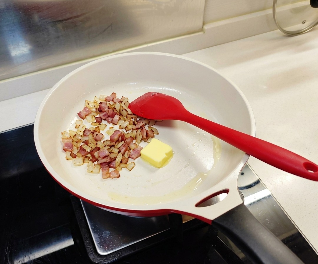 Un cubo de mantequilla amarilla en una sartén blanca junto con el tocino y la cebolla ya cocinados.