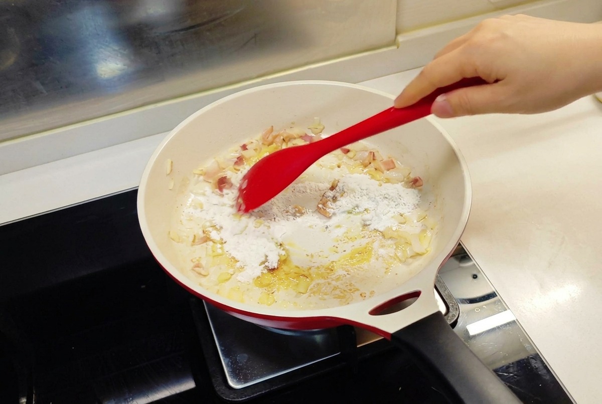 Una espátula roja integrando la harina en una sartén que contiene cebolla sofrita y mantequilla derretida.