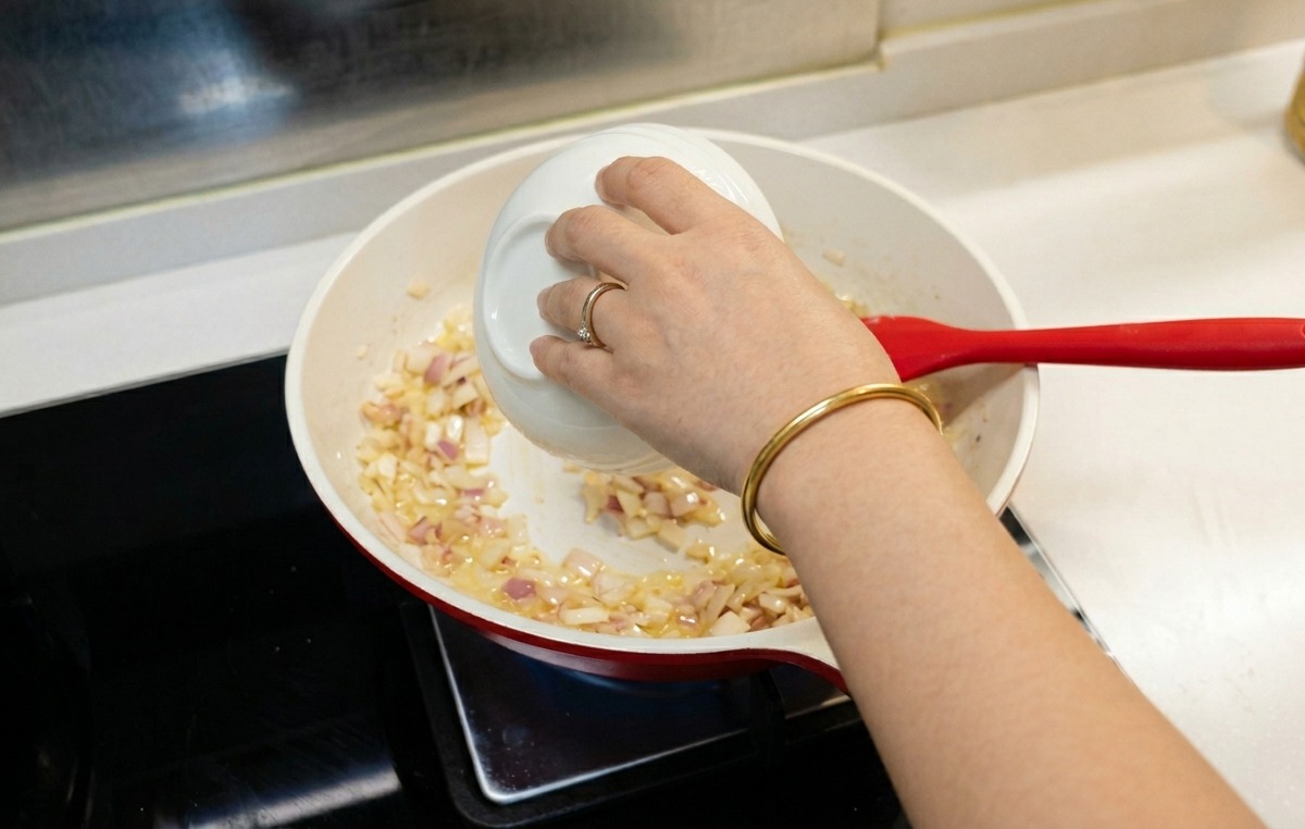 Una persona vertiendo agua desde un pequeño cuenco blanco en una sartén con cebollas cocinadas y harina.