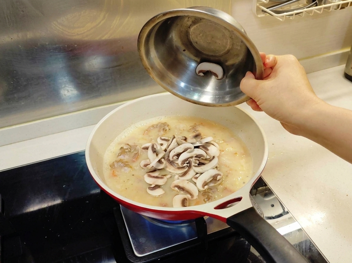 Láminas de champiñones frescos volcándose desde un cuenco metálico a una sartén con una base de salsa cremosa y camarones.