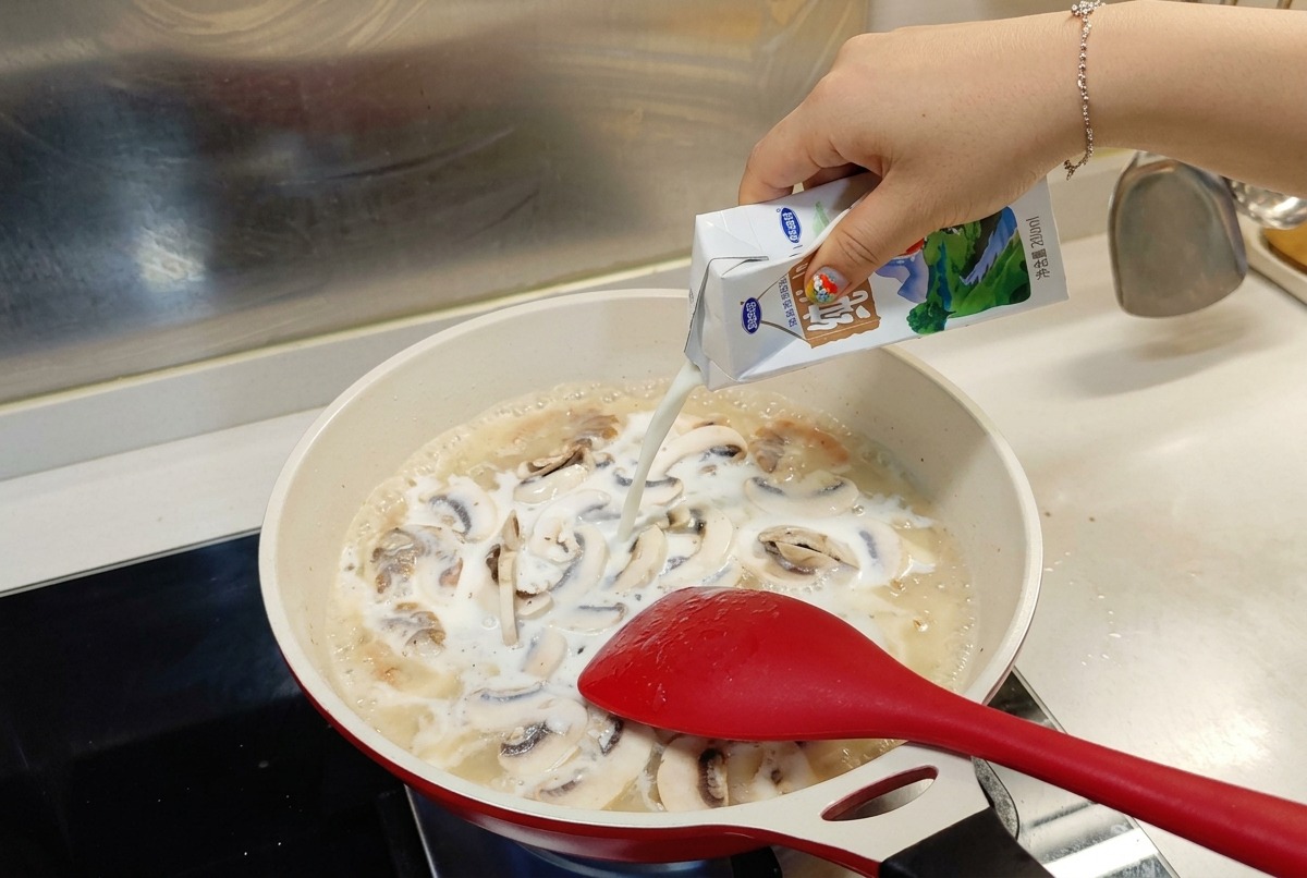 Leche vertiéndose desde un brik blanco y azul en una sartén con camarones y champiñones cocinándose a fuego lento.