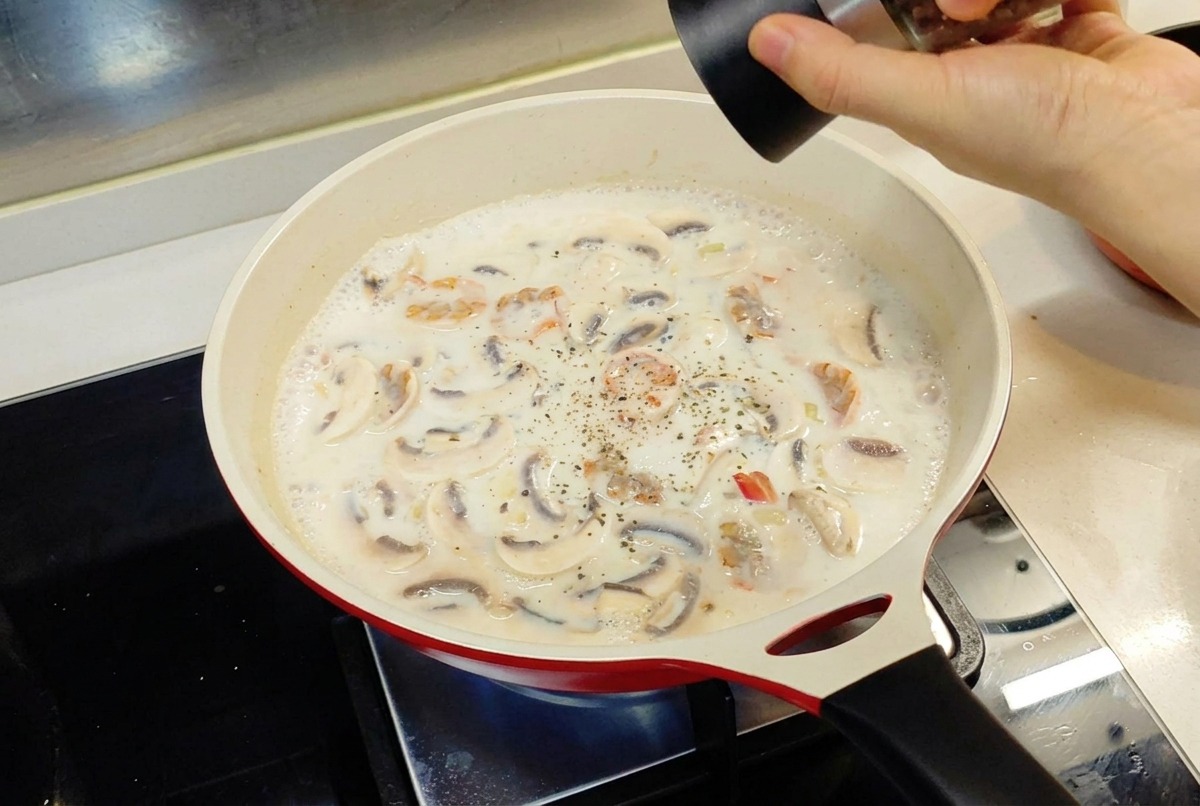 Una mano utilizando un molinillo de pimienta para sazonar una salsa blanca que hierve a fuego lento en una sartén roja.