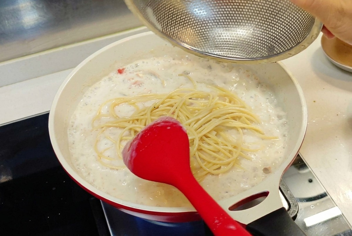 Espaguetis cocinados trasladándose desde un colador de malla hacia la sartén rebosante de salsa blanca.