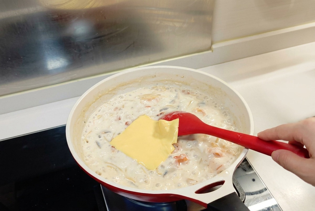 Una rebanada cuadrada de queso amarillo colocada sobre la mezcla de pasta y salsa blanca en una sartén con una cuchara roja.