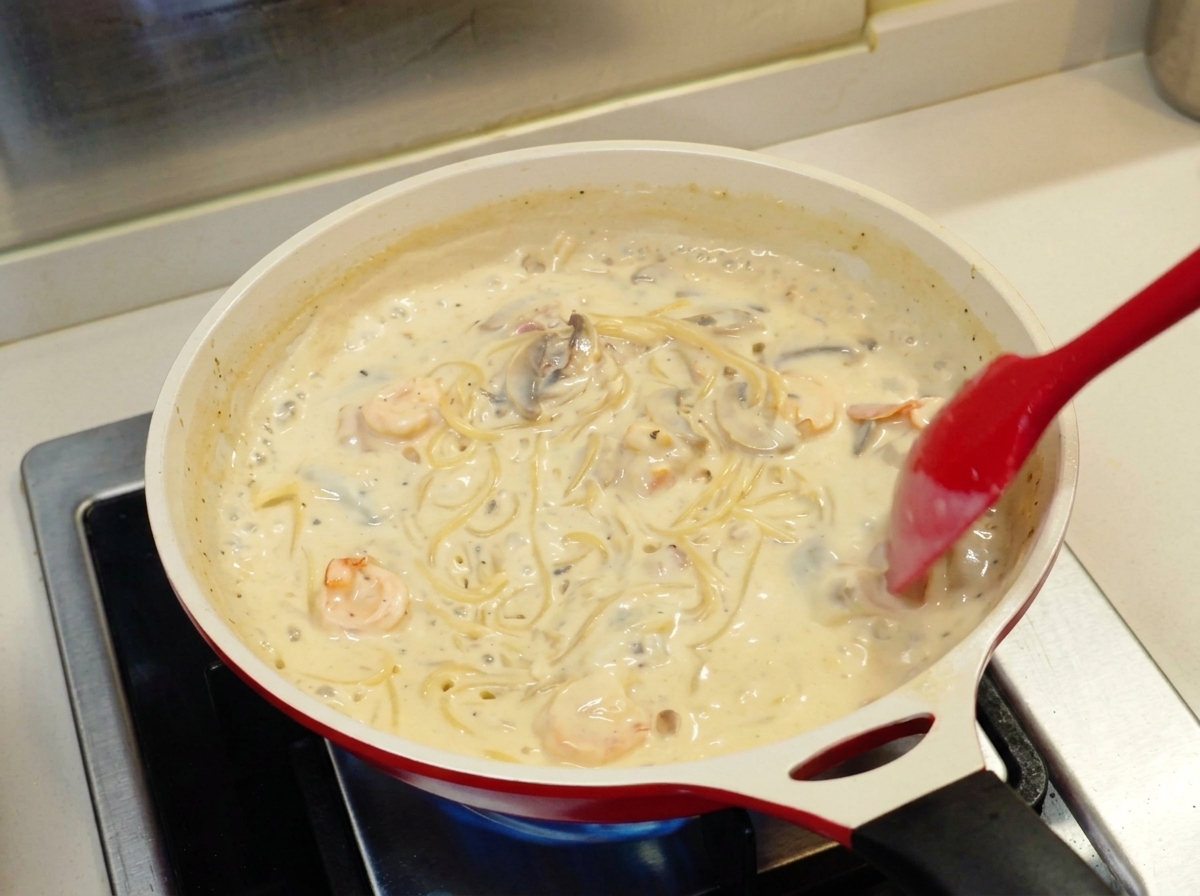 Una espátula roja mezclando espaguetis en una salsa blanca espesa con camarones y champiñones dentro de una sartén cerámica blanca.