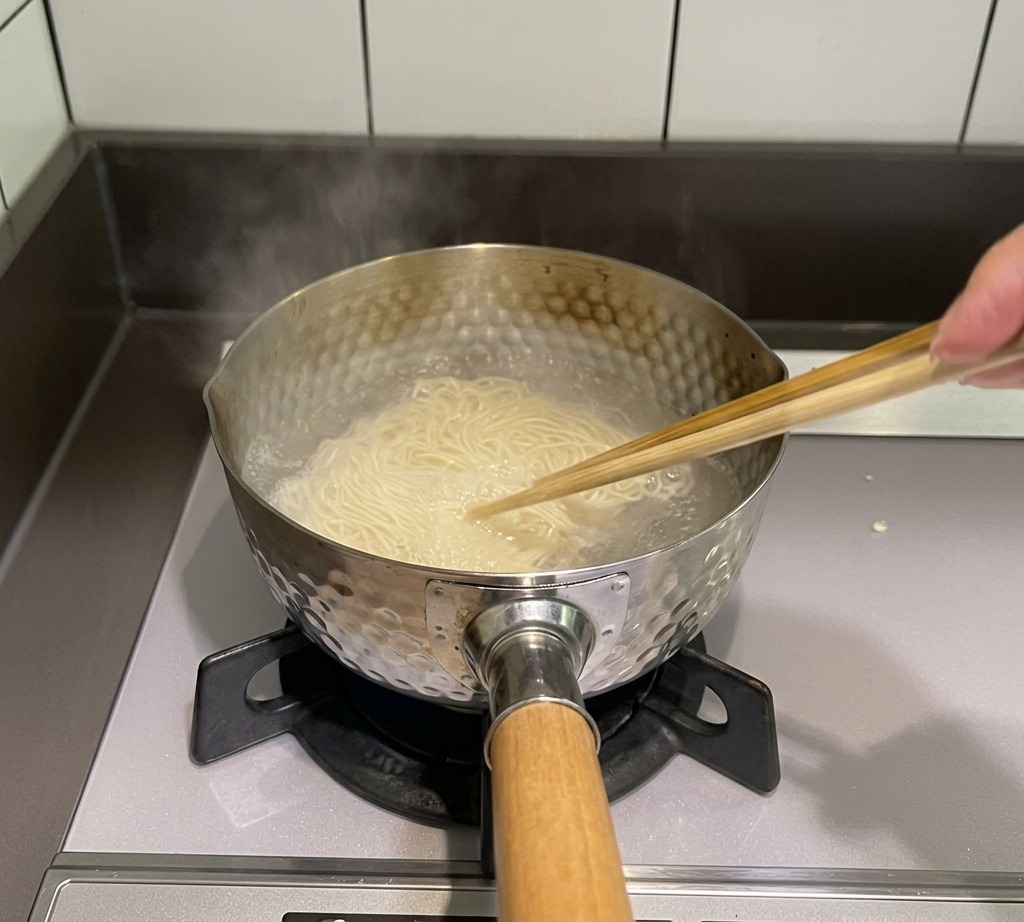 Un par de palillos de madera removiendo fideos finos en una olla con agua hirviendo sobre la estufa.