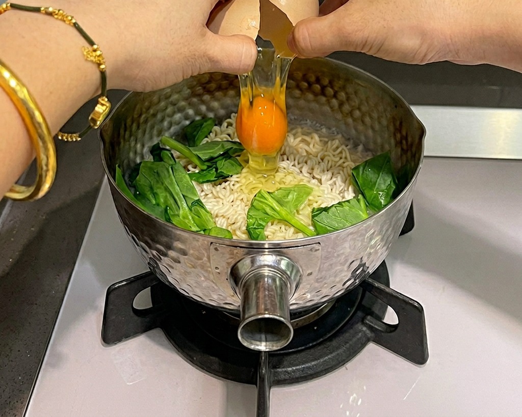 Dos manos cascando un huevo crudo sobre una olla de fideos instantáneos hirviendo con espinacas verdes.