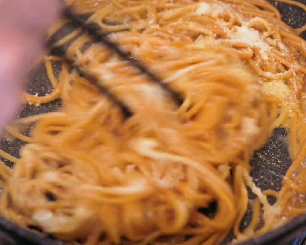 Toma de acción de pasta integral siendo removida firmemente con palillos negros en una sartén, mostrando el queso en polvo visible y la mantequilla derritiéndose para formar una salsa.