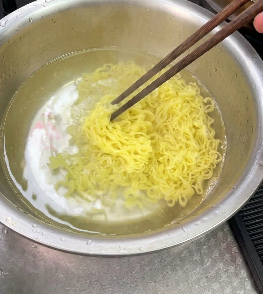 Fideos amarillos cocidos siendo enjuagados y mezclados con palillos en un tazón de acero inoxidable lleno de agua fría.