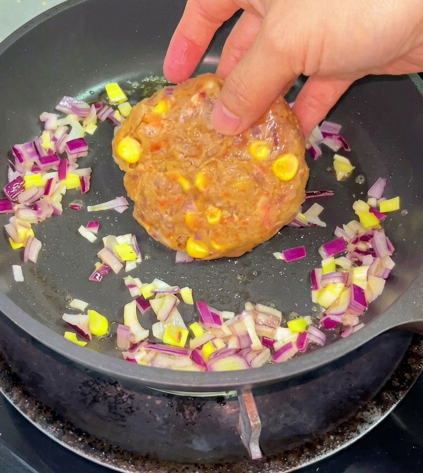 Una mano colocando una porción de carne de res cruda con granos de maíz en una sartén con cebolla morada picada y mantequilla derretida.