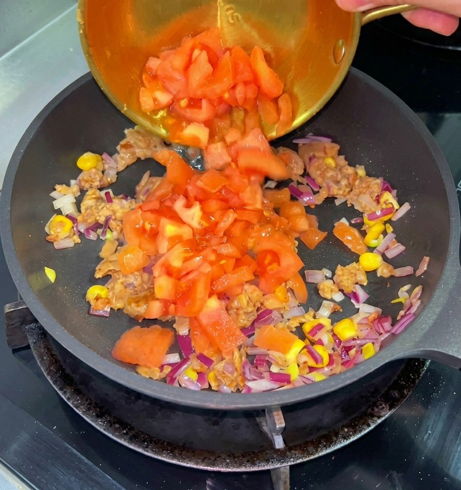 Tomates rojos picados cayendo desde un pequeño tazón de latón a una sartén con carne cocida, maíz y cebolla morada.