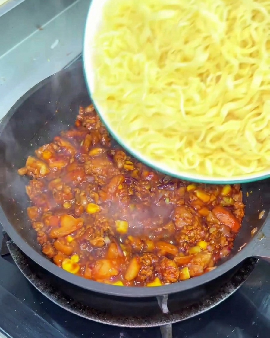 Un tazón de fideos amarillos rizados vertiéndose en una sartén llena de una salsa espesa de tomate de color rojo oscuro con carne.