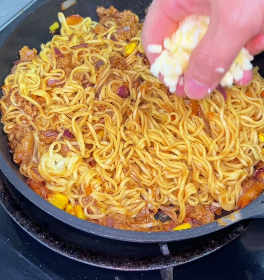 Un primer plano de una mano esparciendo queso blanco rallado sobre una sartén de fideos salteados mezclados con carne molida, cebolla y maíz.