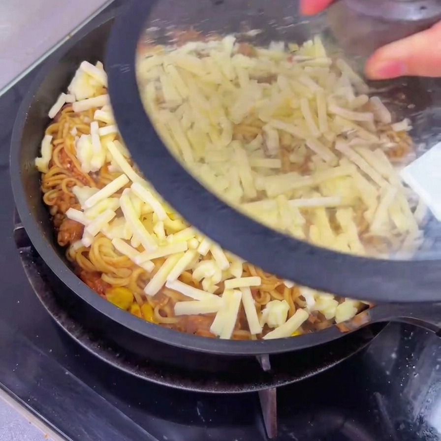 Una persona colocando una tapa de vidrio transparente en una sartén grande que contiene fideos con carne cubiertos con una capa de queso rallado sin derretir.