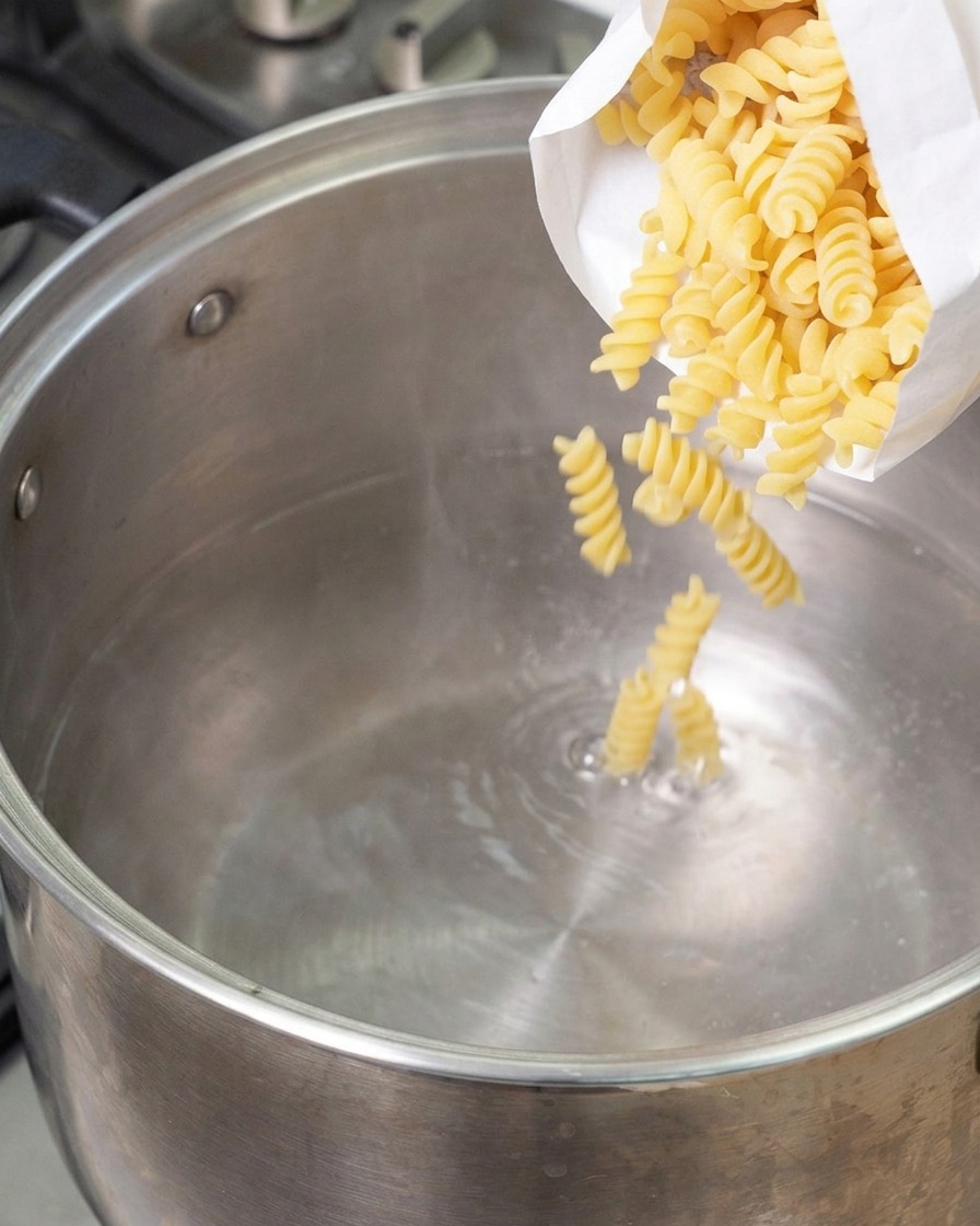 Pasta fusilli seca en espiral cayendo desde una bolsa blanca dentro de una olla grande de acero inoxidable con agua hirviendo.