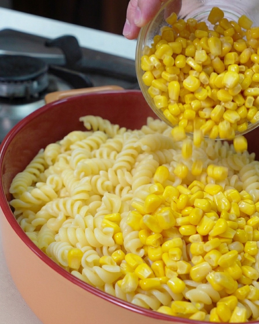 Un bol de vidrio vertiendo granos de maíz dulce de color amarillo brillante sobre una base de fusilli cocidos en una fuente para horno redonda.