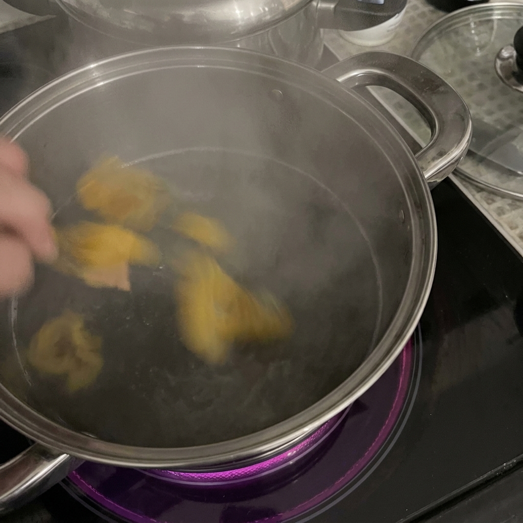 Una mano añadiendo pasta rigatoni de color amarillo a una olla de acero inoxidable con agua hirviendo sobre una placa de inducción.