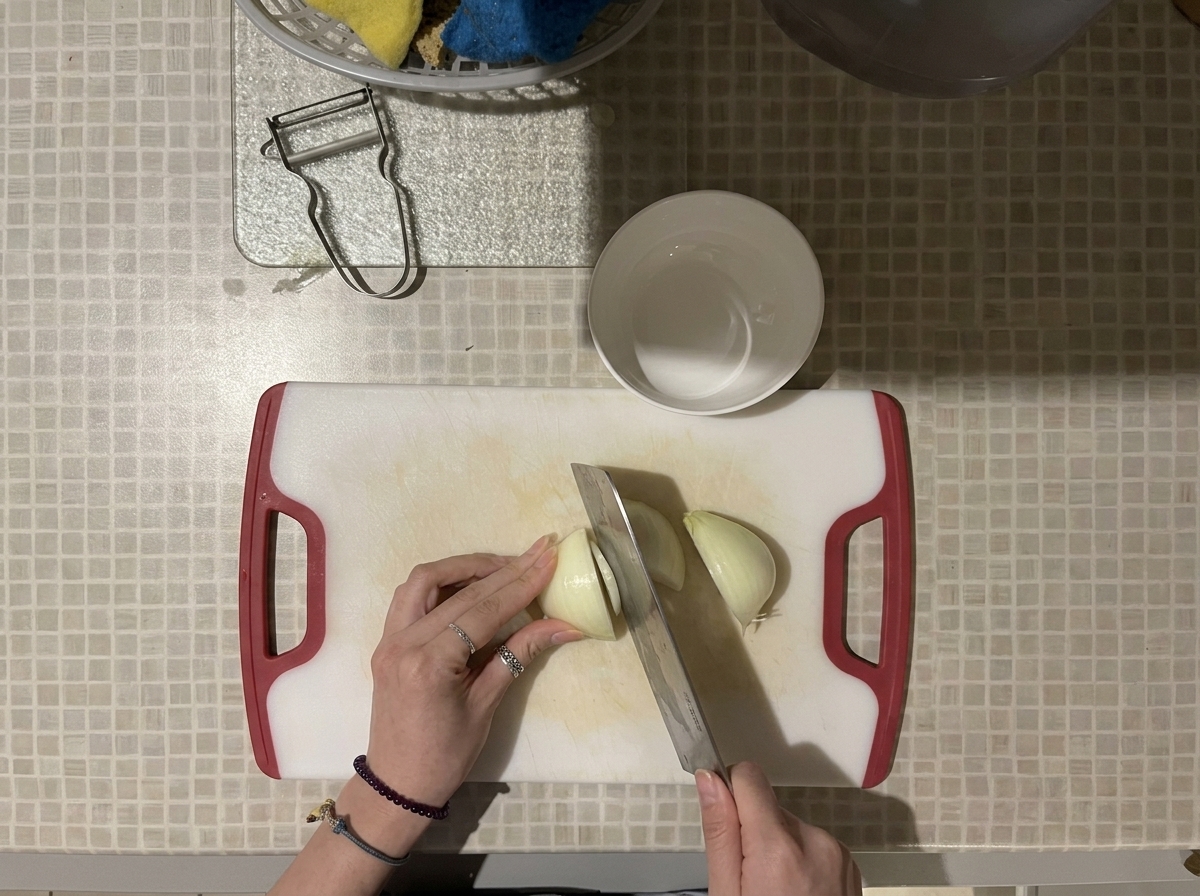 Vista cenital de unas manos cortando una cebolla blanca en cubos pequeños con un cuchillo grande sobre una tabla de cortar blanca con mangos rojos.