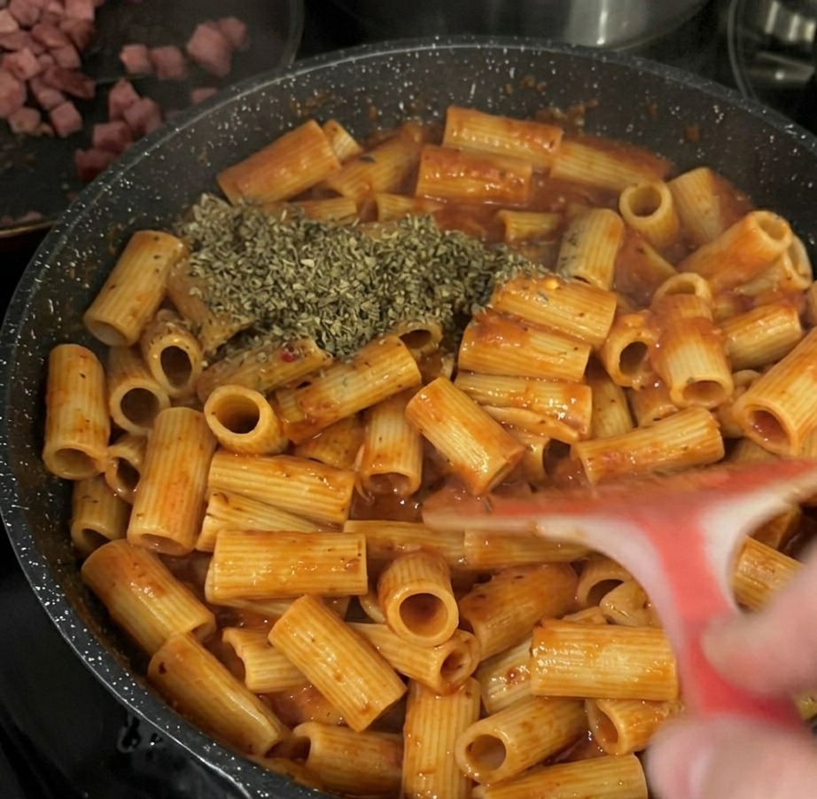 Una cantidad generosa de hojas de albahaca seca espolvoreadas sobre la pasta rigatoni con salsa de tomate dentro de una sartén negra.