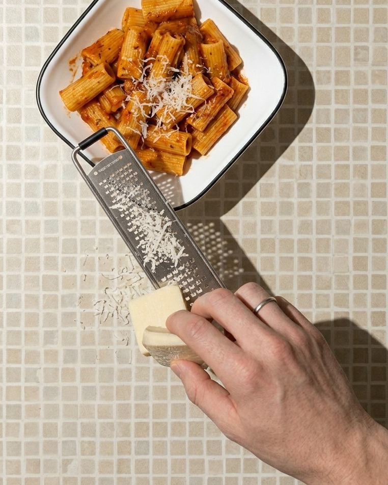 Primer plano de una mano rallando un bloque de queso blanco sobre un plato cuadrado blanco lleno de pasta rigatoni con salsa roja.
