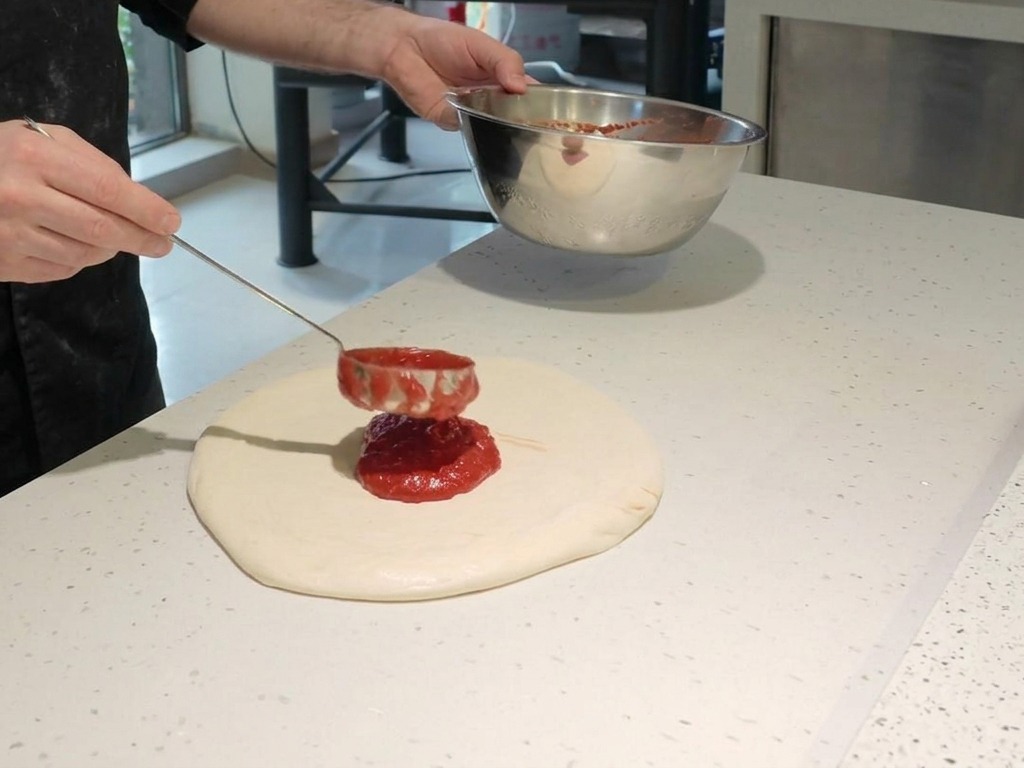 Un chef usando un cucharón metálico para esparcir salsa de tomate roja en espiral sobre la masa de pizza cruda.