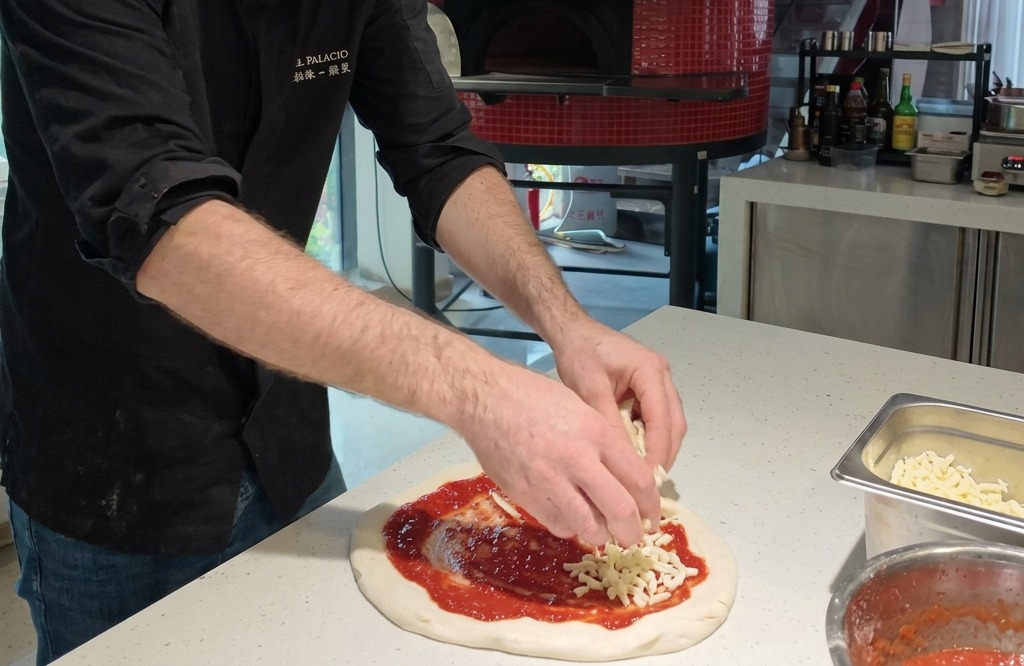 Manos distribuyendo trozos de queso mozzarella fresco sobre una base de pizza cubierta de salsa de tomate.