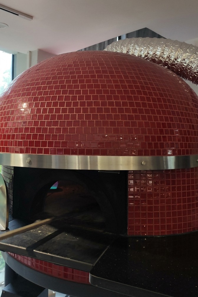 Un horno de leña con exterior de azulejos rojos mientras se introduce una pizza con una pala metálica larga.