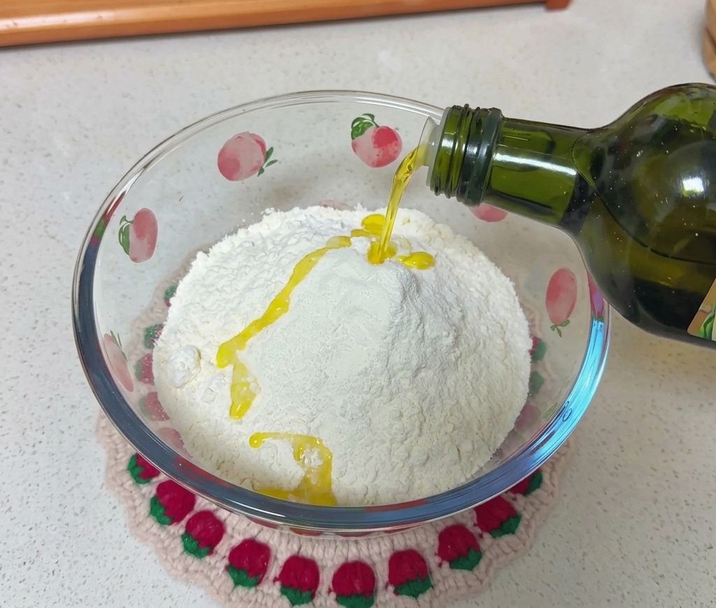 Aceite de oliva cayendo desde una botella de vidrio verde oscuro sobre una montaña de harina blanca dentro de un bol de vidrio decorado con motivos de melocotón.