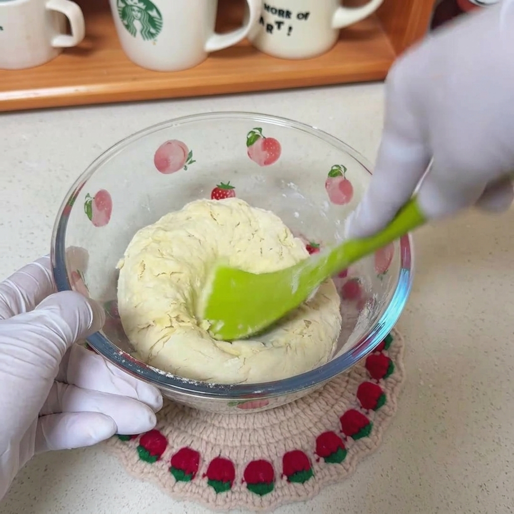 Una espátula verde mezclando una masa rústica de harina, agua y aceite en un bol de vidrio transparente decorado con frutas.