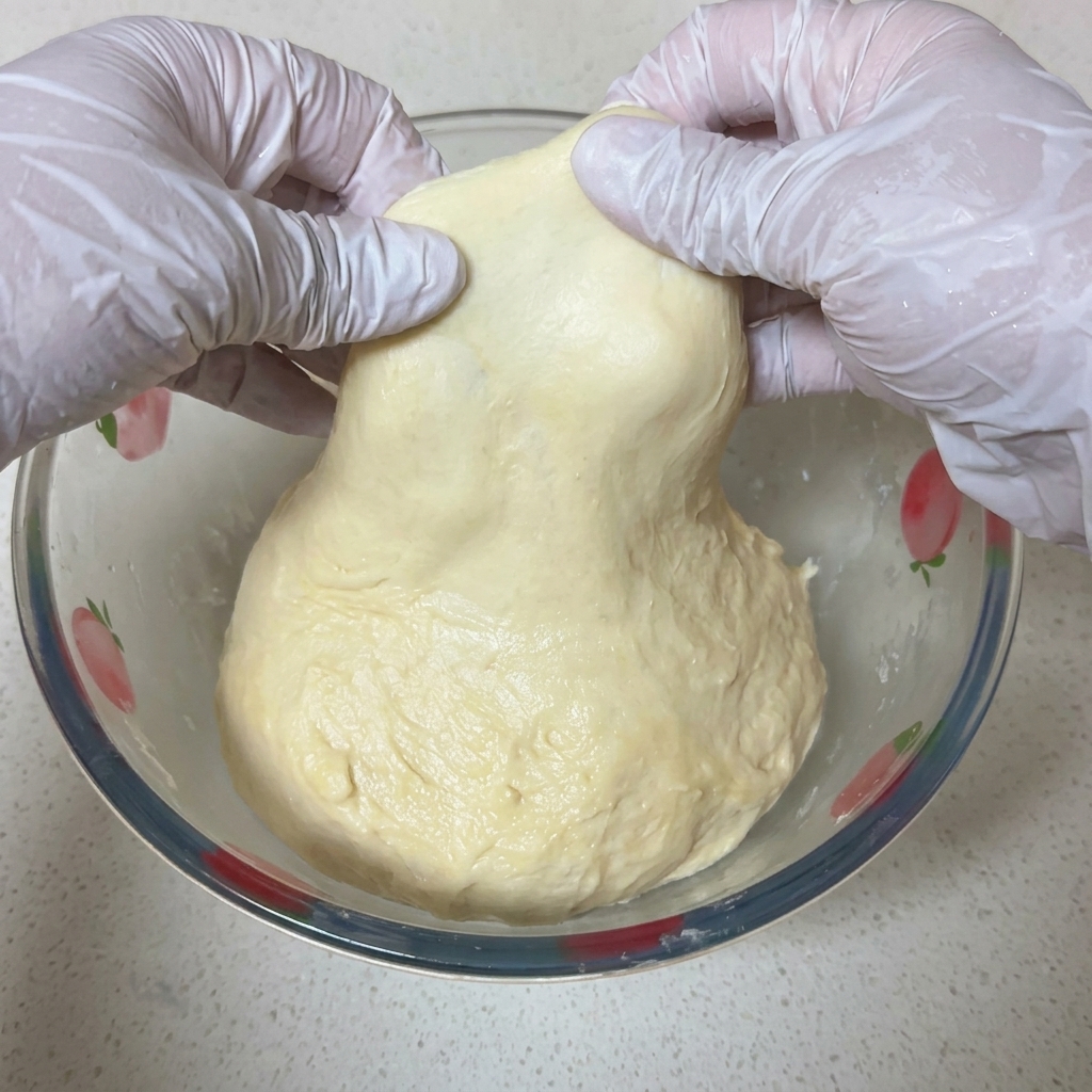 Dos manos con guantes blancos estirando delicadamente un trozo de masa de pizza para mostrar una membrana fina y translúcida a contraluz.