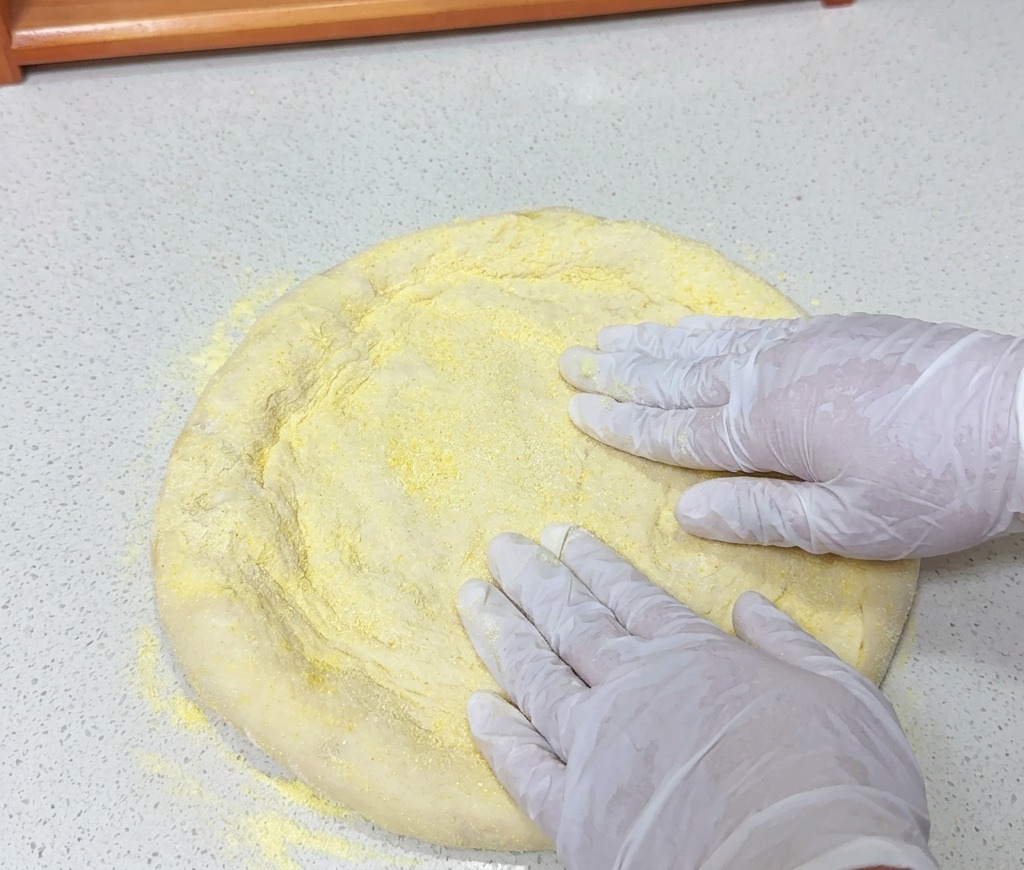 Manos con guantes blancos presionando una pieza redonda de masa de pizza cubierta con sémola de maíz amarilla sobre una encimera clara.