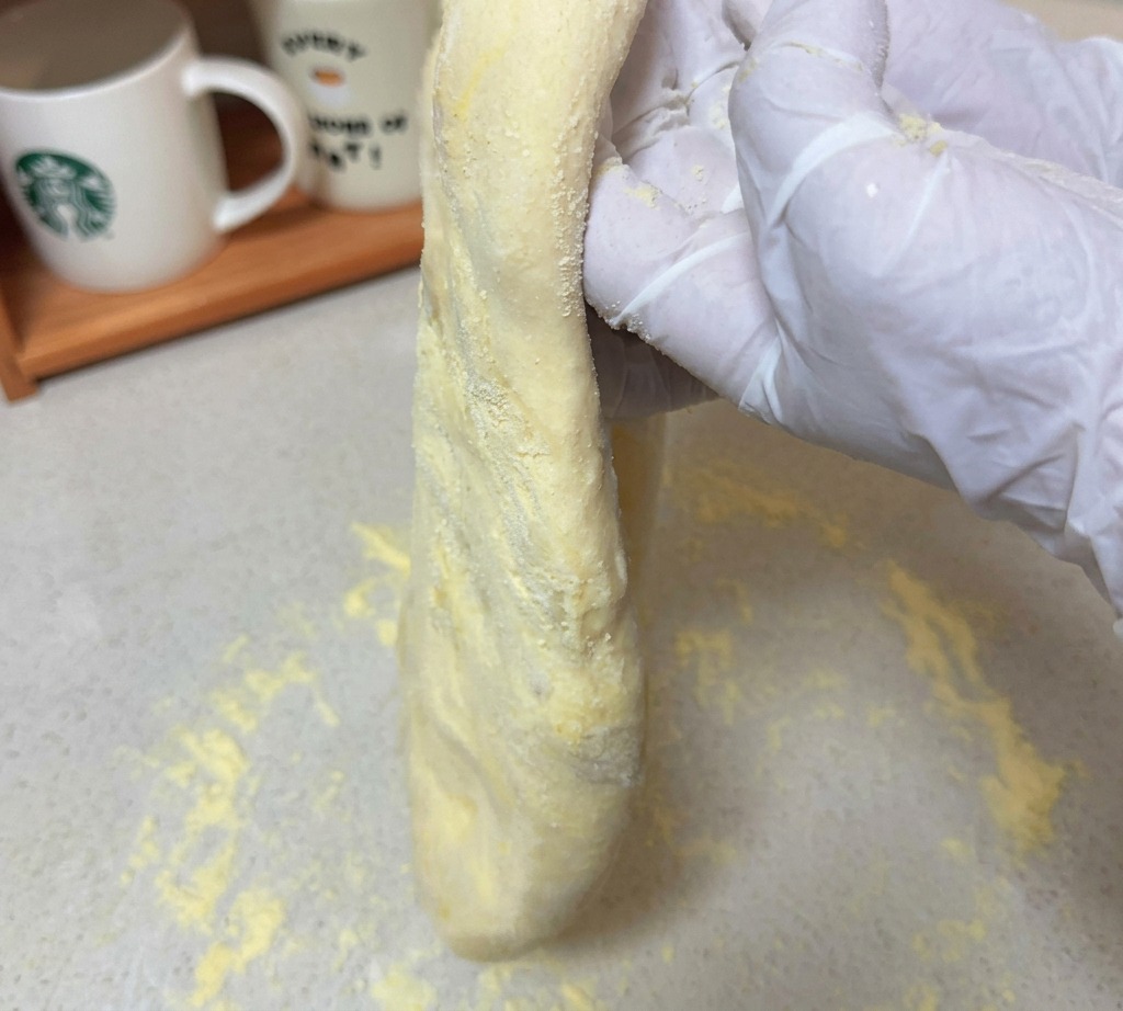 Una porción de masa de pizza estirada sostenida verticalmente por manos con guantes sobre una superficie enharinada.