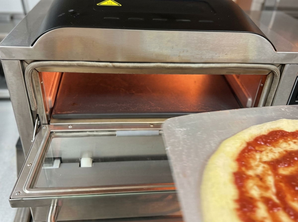 Una pizza cruda con salsa de tomate y mozzarella siendo introducida desde una pala metálica al interior iluminado de un horno eléctrico profesional para pizzas.