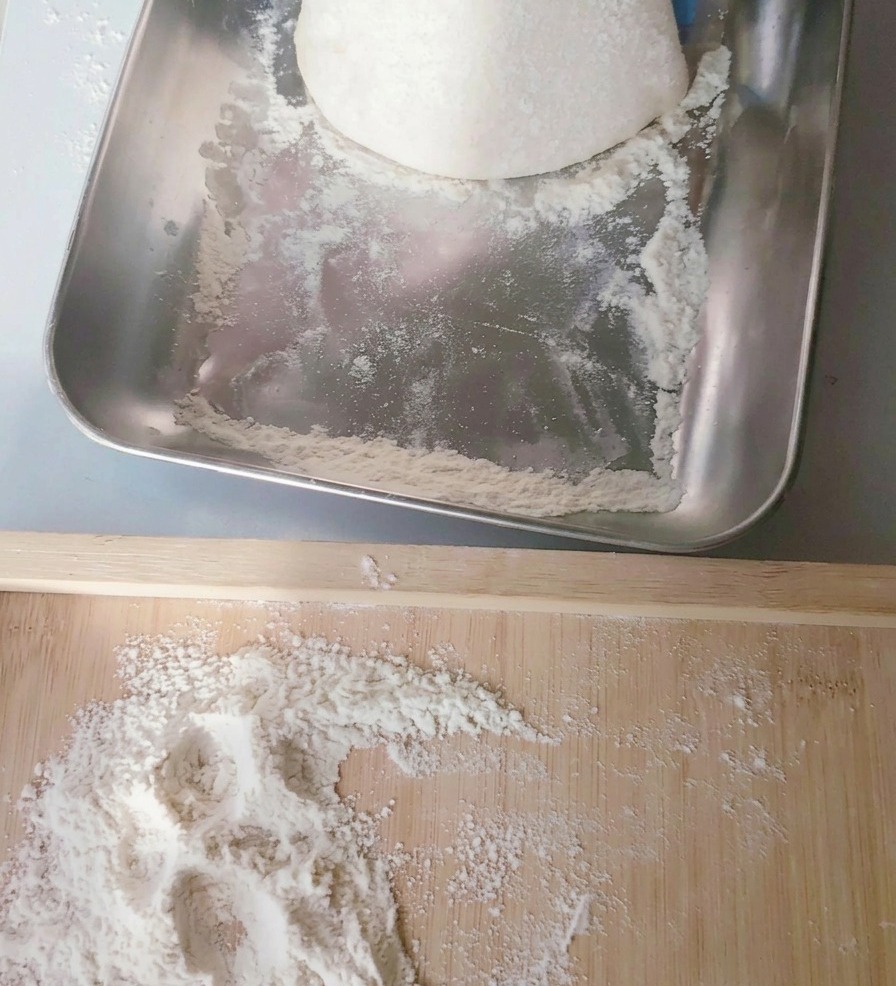 Una bola redonda de masa de pizza pálida que se levanta de una bandeja de acero inoxidable hacia una tabla de madera espolvoreada con harina.